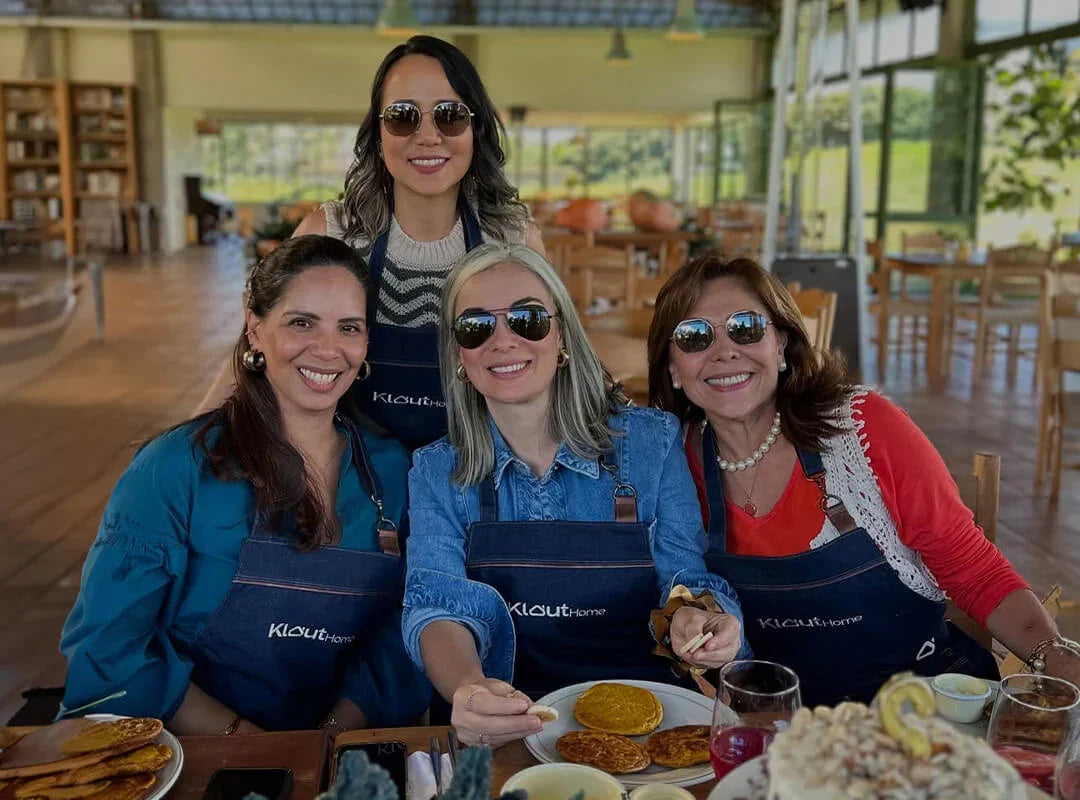 Mujeres sonrientes con delantales sentadas en mesa de desayuno en cocina luminosa, panqueques y bebidas